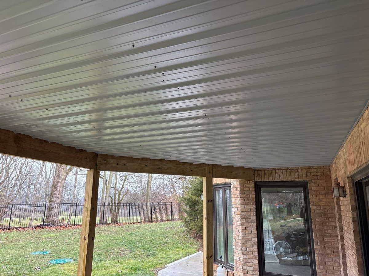 White corrugated metal ceiling installed under a wooden deck structure next to a brick house.