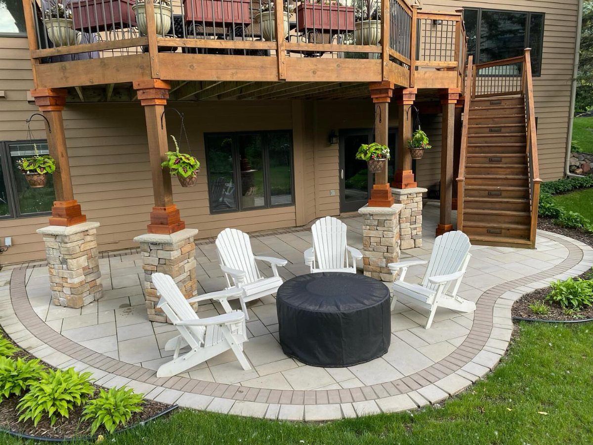 White Adirondack chairs and a black ottoman arranged on a stone patio under a wooden deck.
