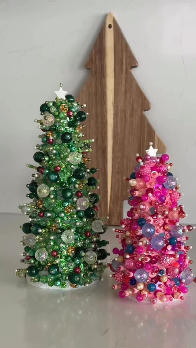Two small beaded Christmas trees, one green and one pink, sitting on a white surface.