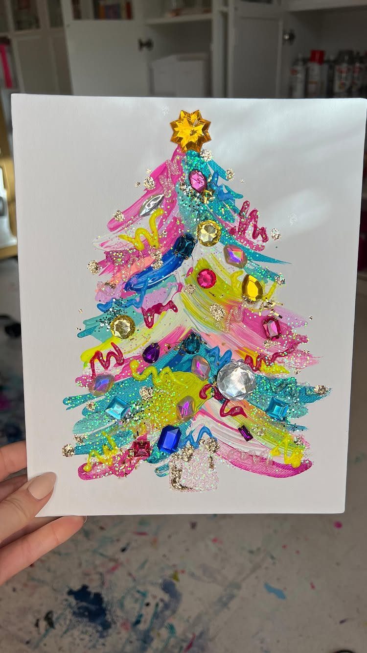 A hand holding a white canvas with a colorful, textured painted Christmas tree decorated with gems.