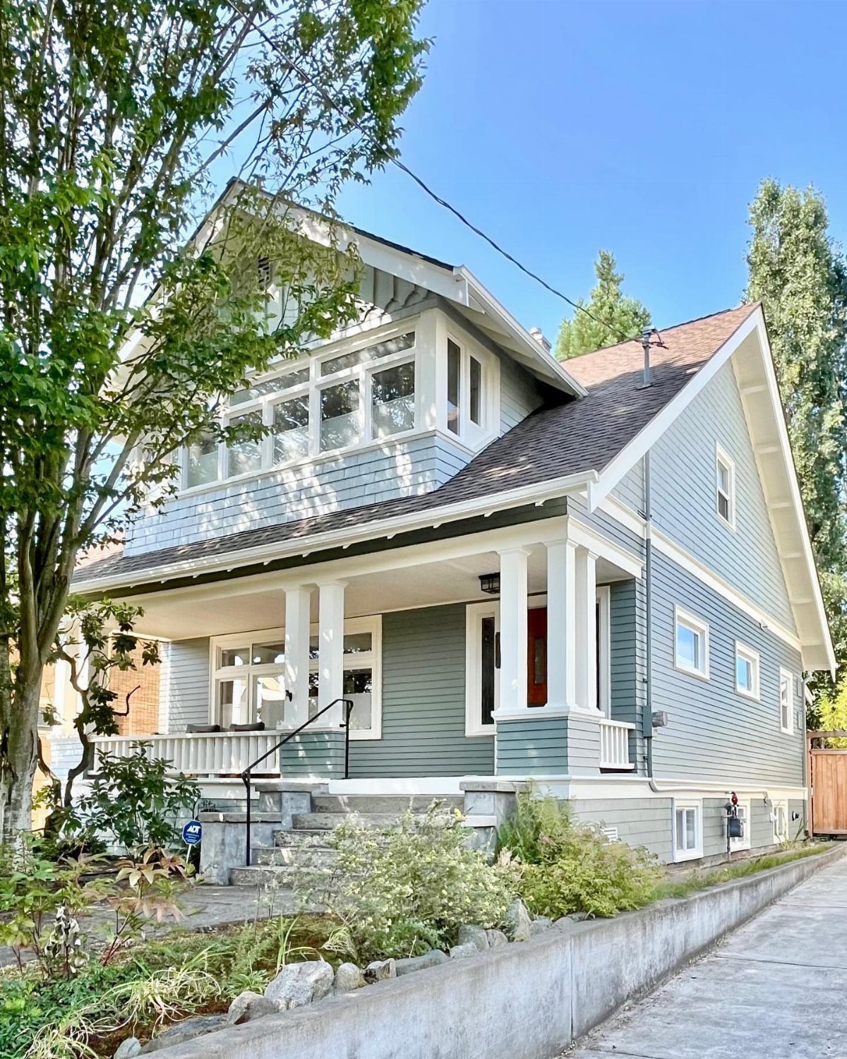 Exterior Color Ideas for 6 Home Styles 3 A blue-grey craftsman house with white trim and a large front porch surrounded by green trees.