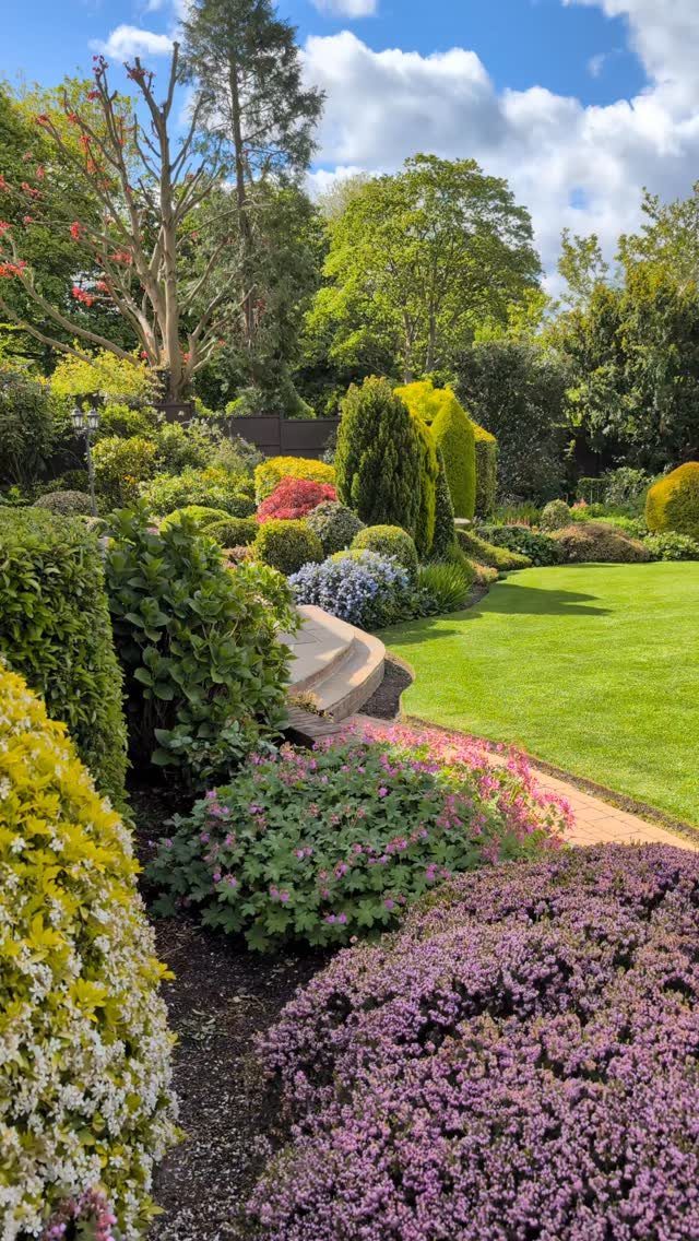 16 Front Lawn Landscaping Ideas for Curb Appeal 10 Lush garden landscape with colorful flowering shrubs, green lawn, and tall trees under a blue sky.