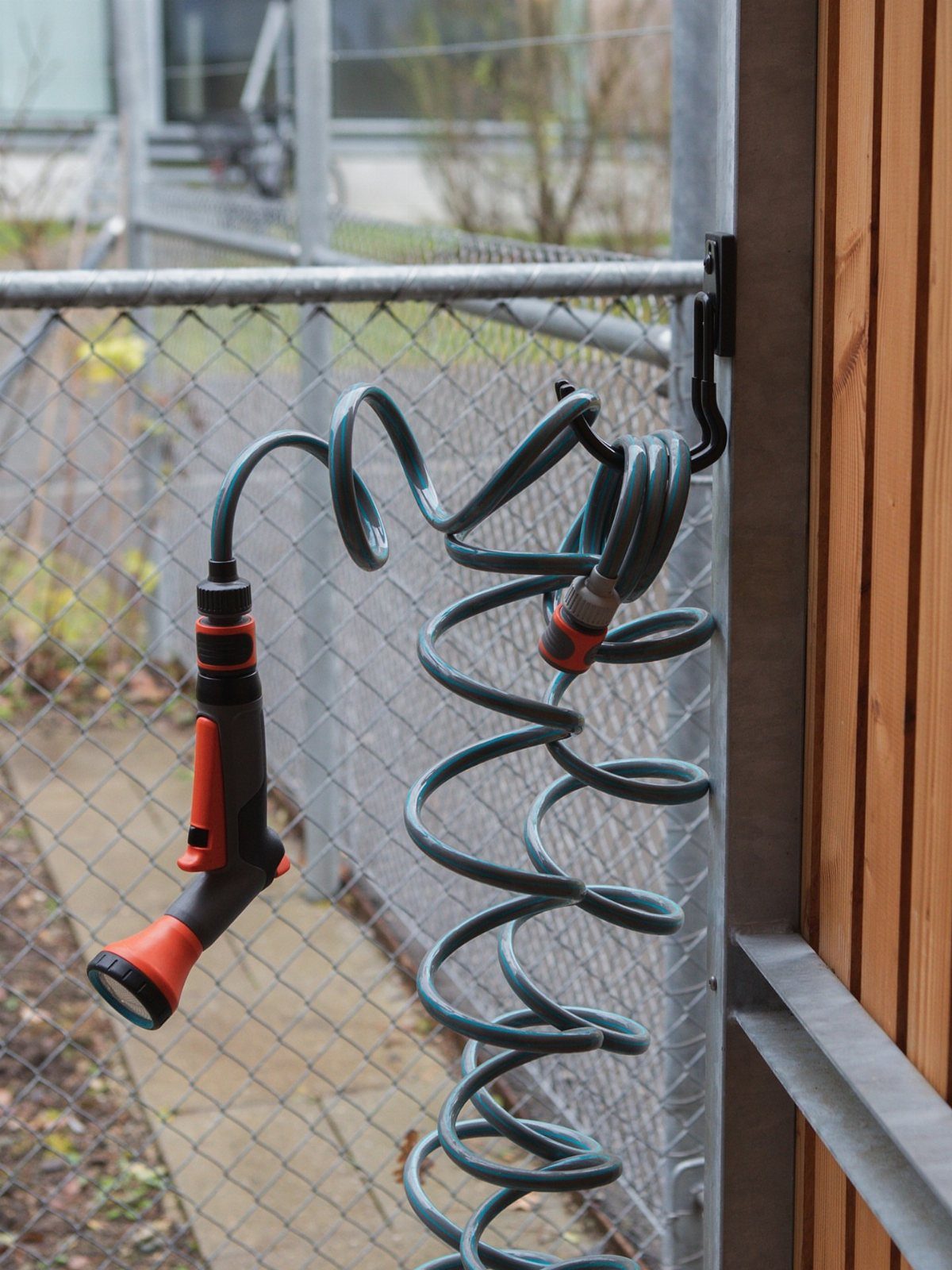 A coiled grey and orange garden hose hanging from a black hook on a metal fence frame.