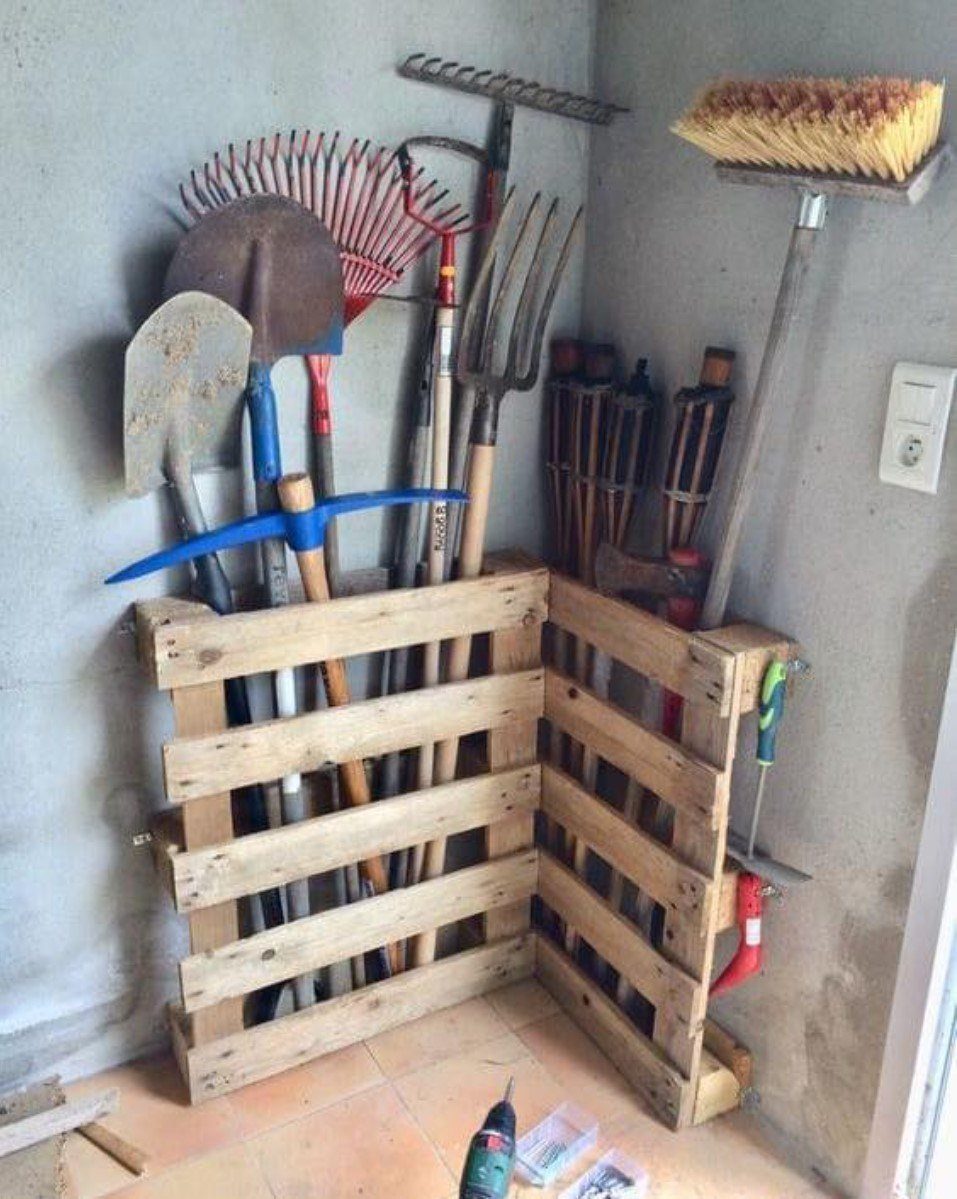 Garden tools stored upright inside a repurposed wooden pallet structure in a corner.