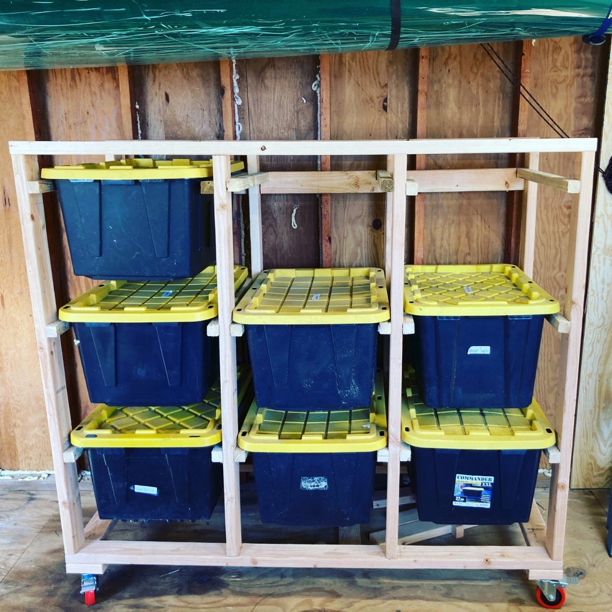 Wooden rolling shelf unit holding several black storage bins with yellow lids inside a shed.