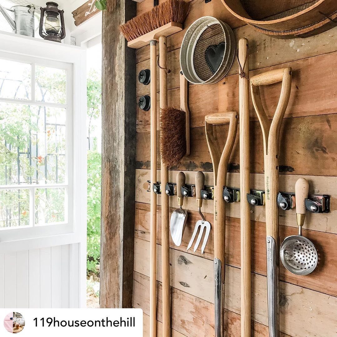 Garden tools like shovels and brushes hanging on a metal rack against a wooden wall.