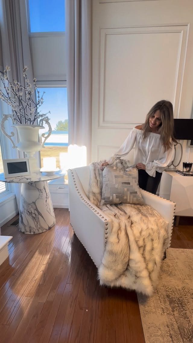 A woman stands by a white armchair covered in a plush faux fur blanket.