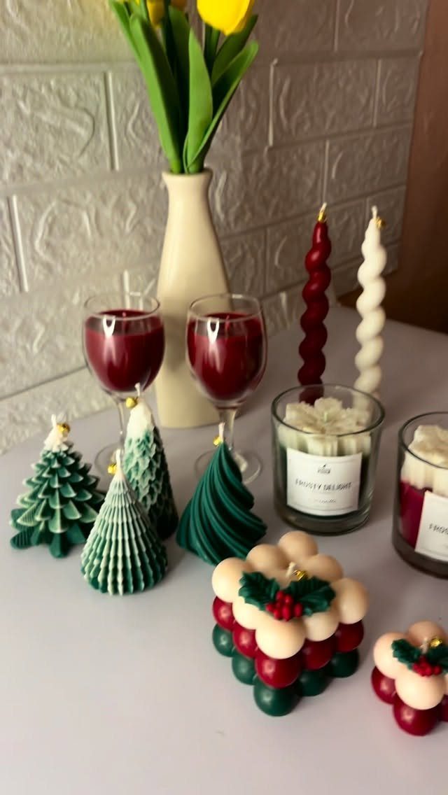 Decorative shaped candles and glasses of red wine on a white table near yellow tulips.