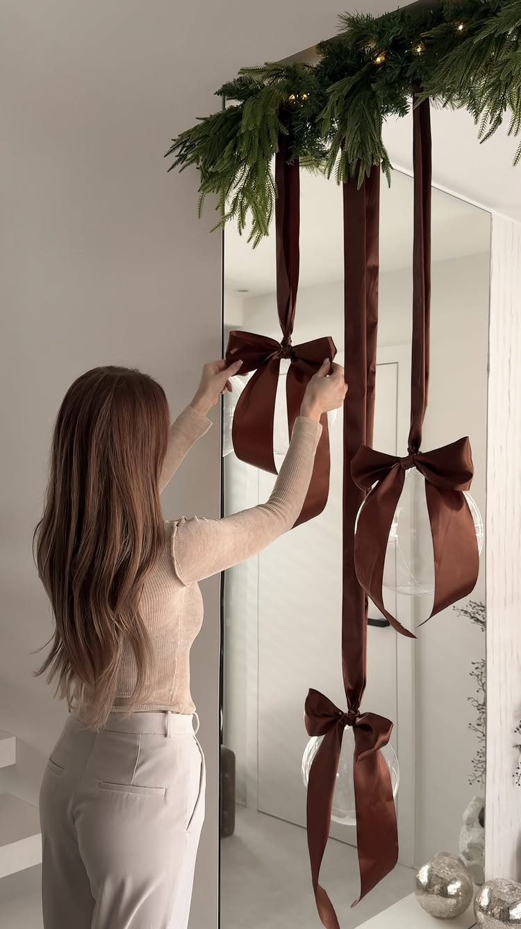 Woman adjusting large brown satin bows hanging from a festive evergreen garland with lights.