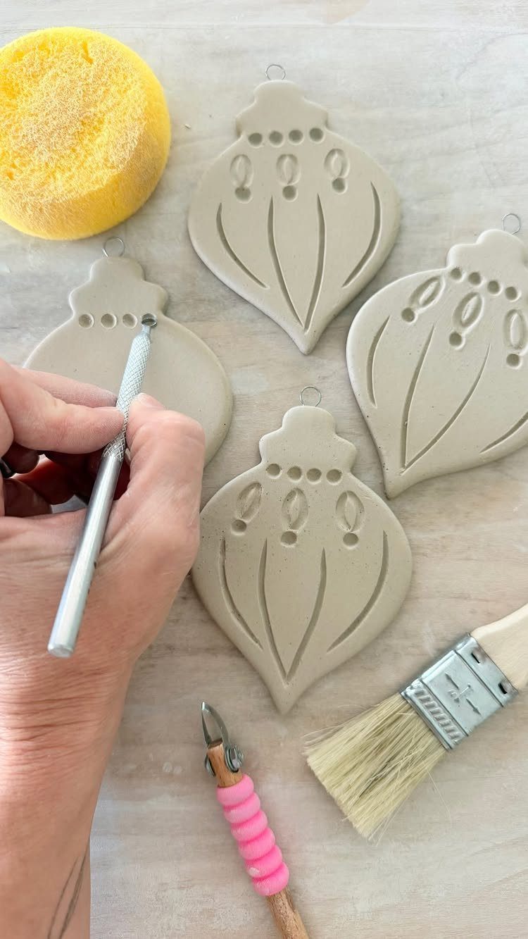 A hand uses a sculpting tool to carve designs into beige clay ornaments on a table.