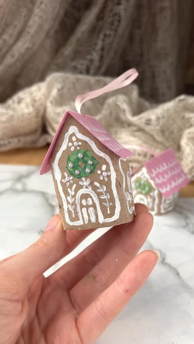 A hand holding a small, decorated gingerbread house Christmas ornament with a pink roof.