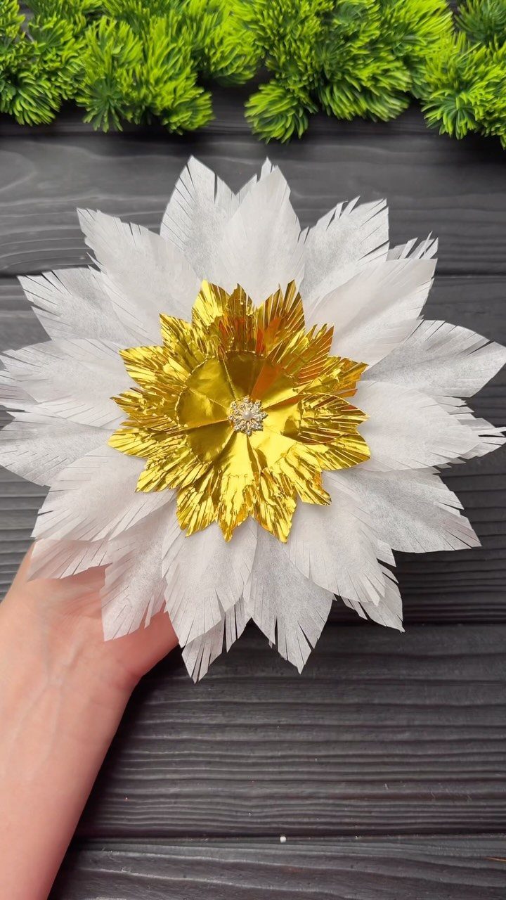 A hand holding a decorative white and gold paper flower ornament against dark wood.
