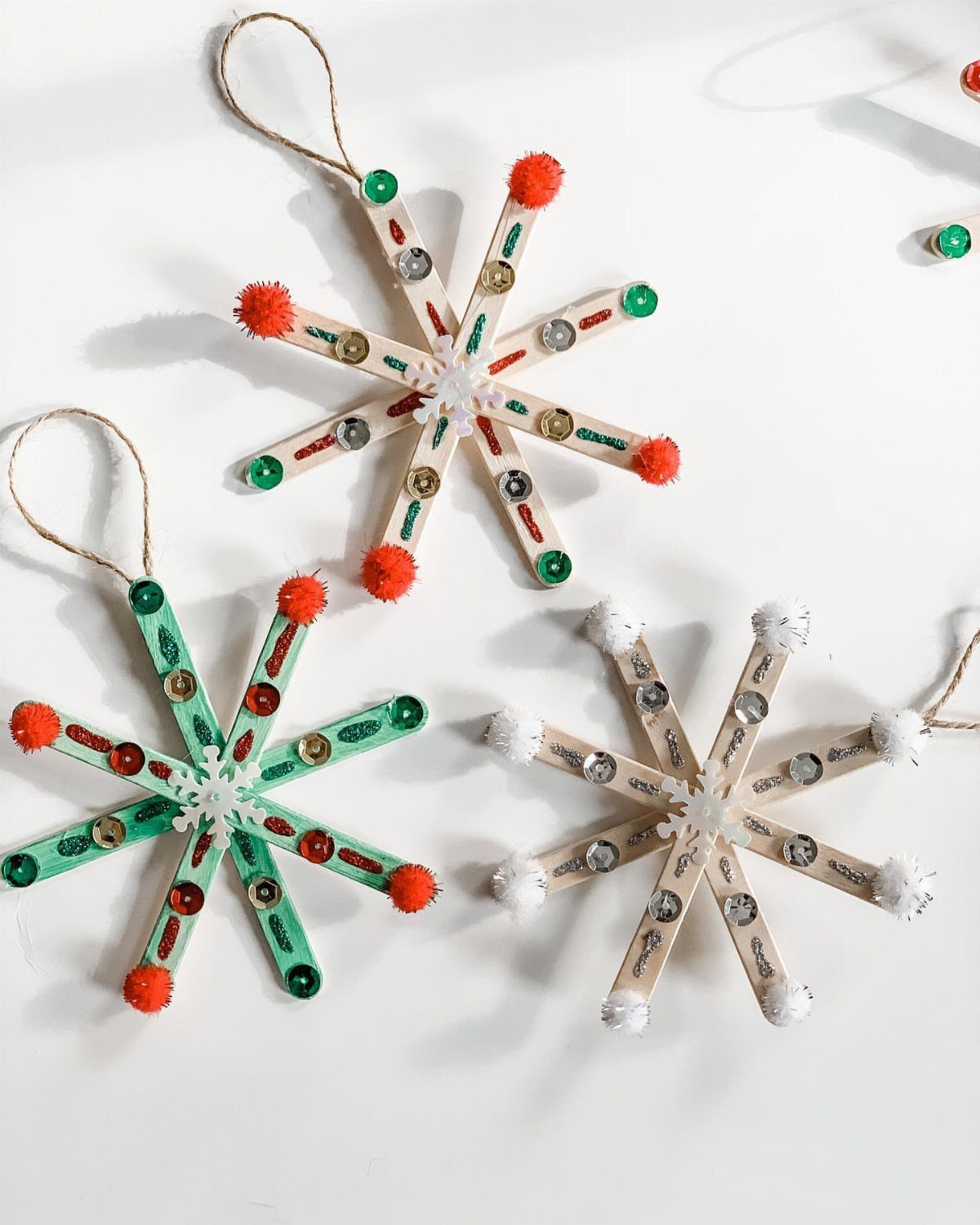 Three handmade snowflake ornaments crafted from colorful popsicle sticks with sequins and pom-poms.