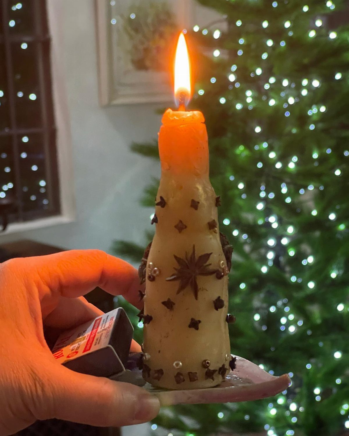 A hand holding a lit, decorated beeswax candle against a blurred Christmas tree background.