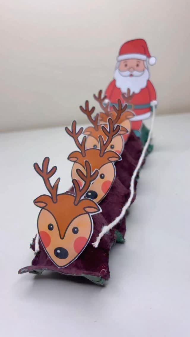 Santa Claus pulling a reindeer sleigh made from a purple egg carton on a white surface.