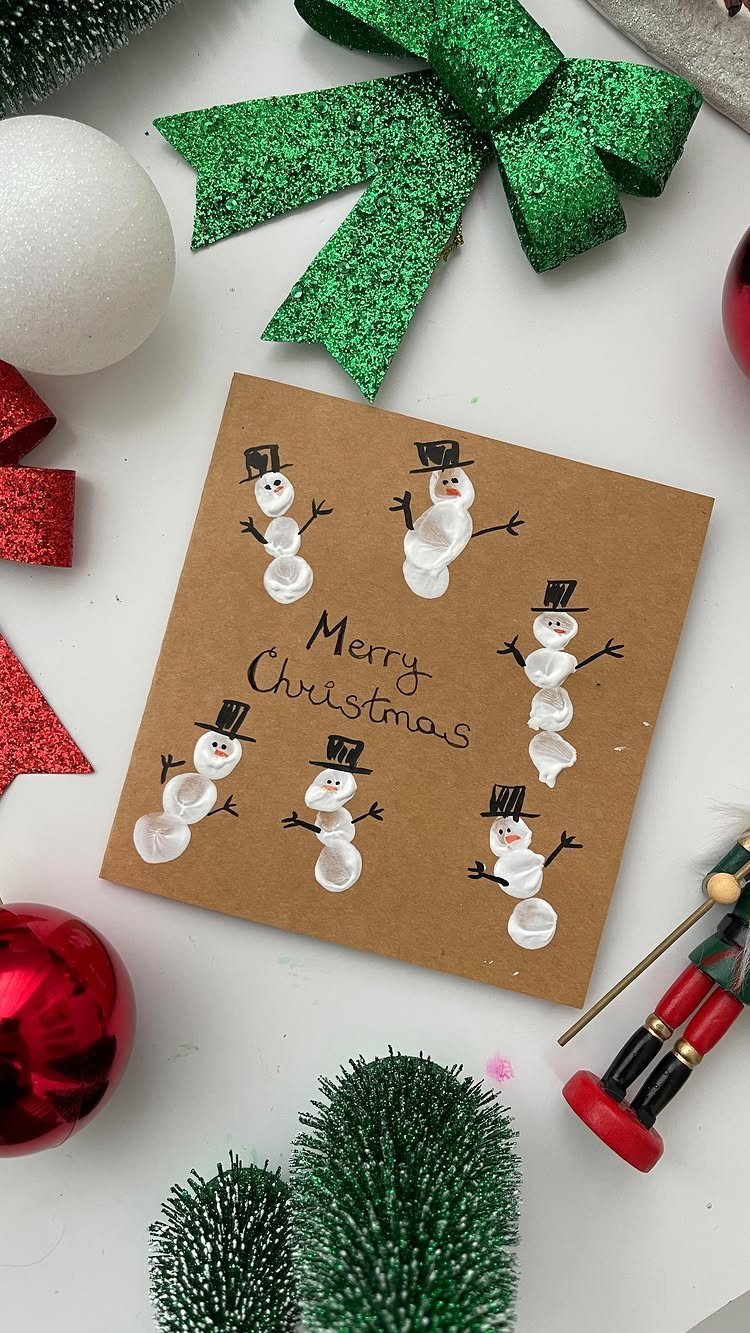 Fingerprint snowmen drawn on a brown card surrounded by festive Christmas decorations and ornaments.