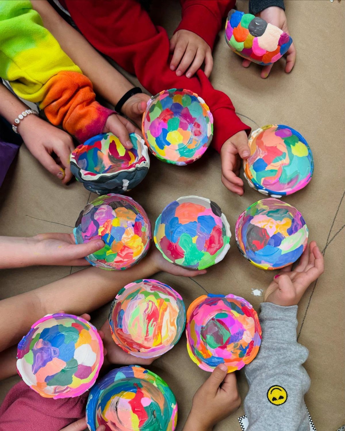 Children holding up various small, brightly hand-painted colorful bowls on a neutral surface.