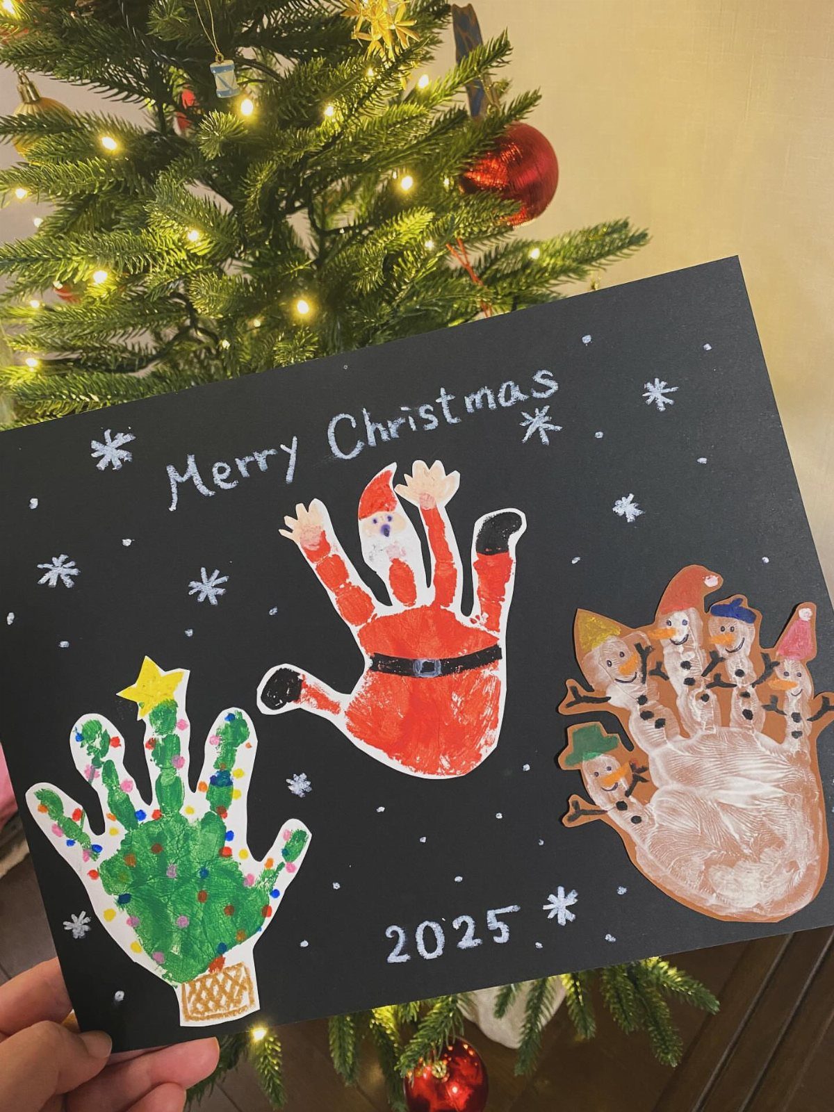 Handprint art on black paper featuring a green tree, Santa, and snowmen near a Christmas tree.