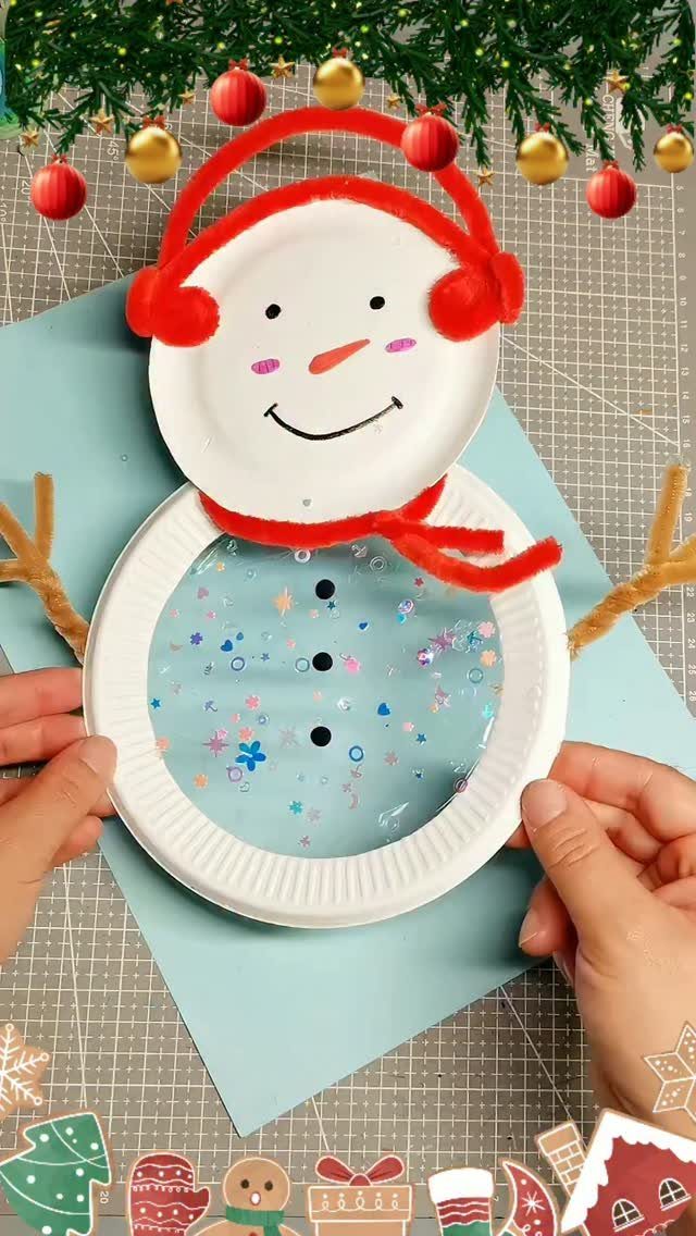 Hands holding a snowman craft made from white paper plates and red pipe cleaners.