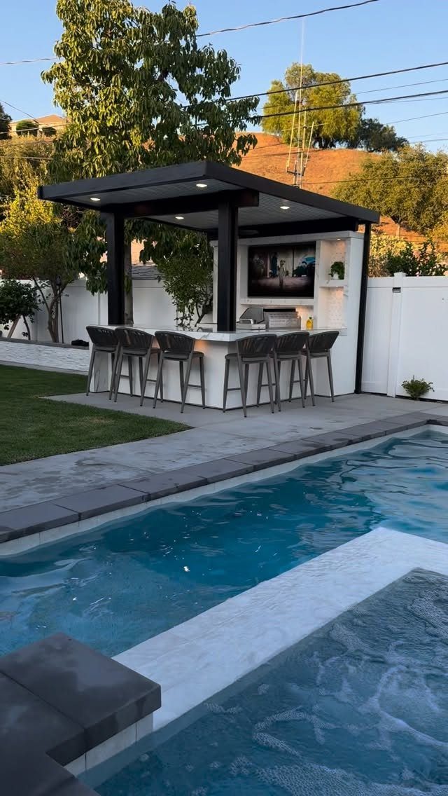 11 Outdoor Kitchen with Pool Ideas That Actually Work 4 Modern outdoor kitchen with bar stools and a television situated next to a swimming pool.