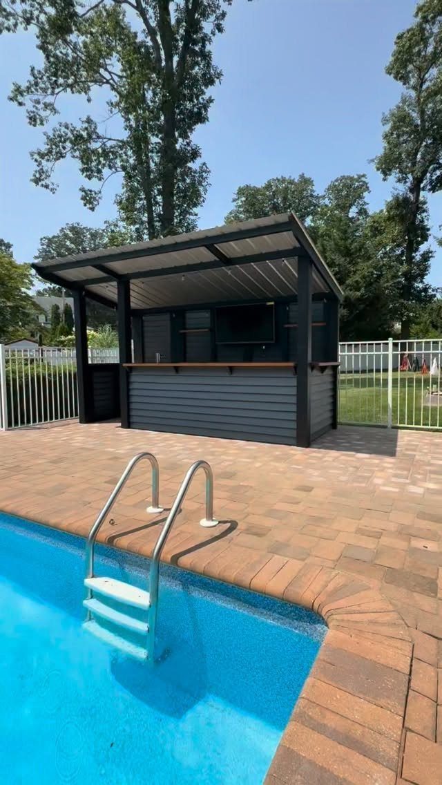 11 Outdoor Kitchen with Pool Ideas That Actually Work 9 A dark grey outdoor bar structure stands on a brick patio beside a blue swimming pool.