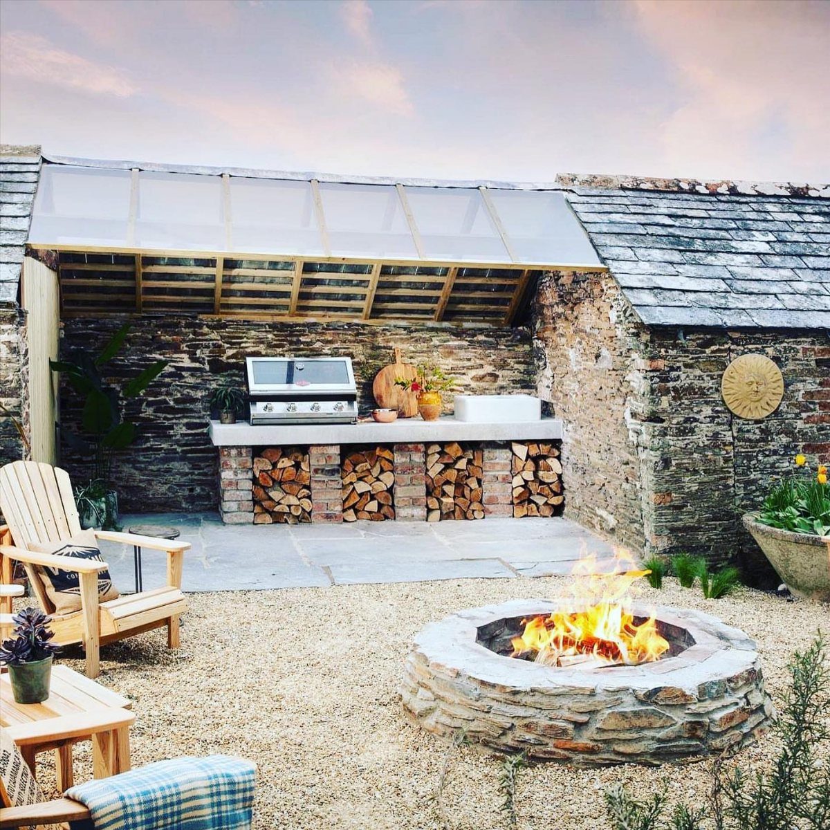 11 Outdoor Kitchen with Pool Ideas That Actually Work 1 Stone outdoor kitchen with a grill and a burning stone fire pit in a gravel courtyard.