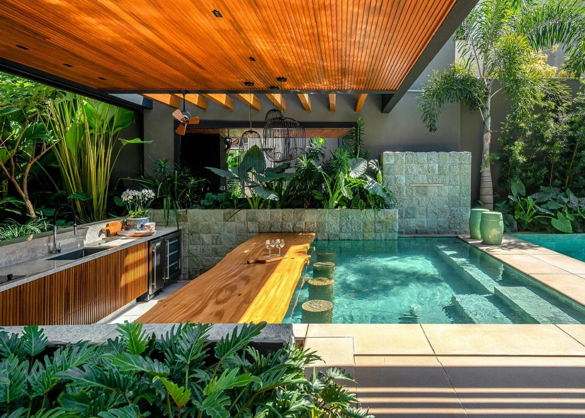 11 Outdoor Kitchen with Pool Ideas That Actually Work 5 Wooden bar counter extending into a turquoise swimming pool with submerged stools and tropical greenery.