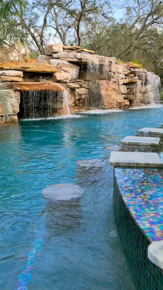 11 Outdoor Kitchen with Pool Ideas That Actually Work 3 Stone waterfalls cascading into a blue swimming pool with stepping stones in the foreground.