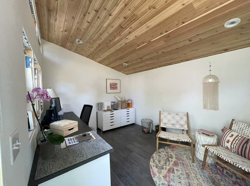 Interior view of a small backyard studio featuring a wooden ceiling, desk, and woven chairs.