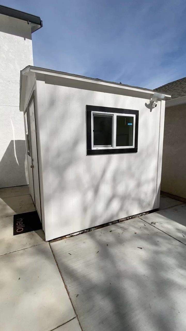 A white backyard studio building with a black window frame sitting on a concrete patio.