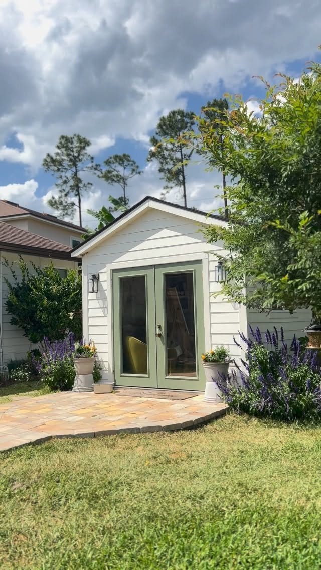 A white wooden garden shed with green French doors sits on a stone patio surrounded by greenery.