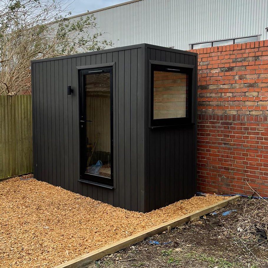 A compact black wooden garden office pod with a glass door and window on wood chips.