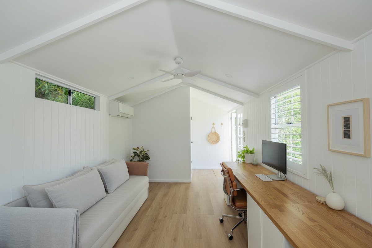 Modern white interior studio with a long wooden desk, computer, grey sofa, and light wood floors.