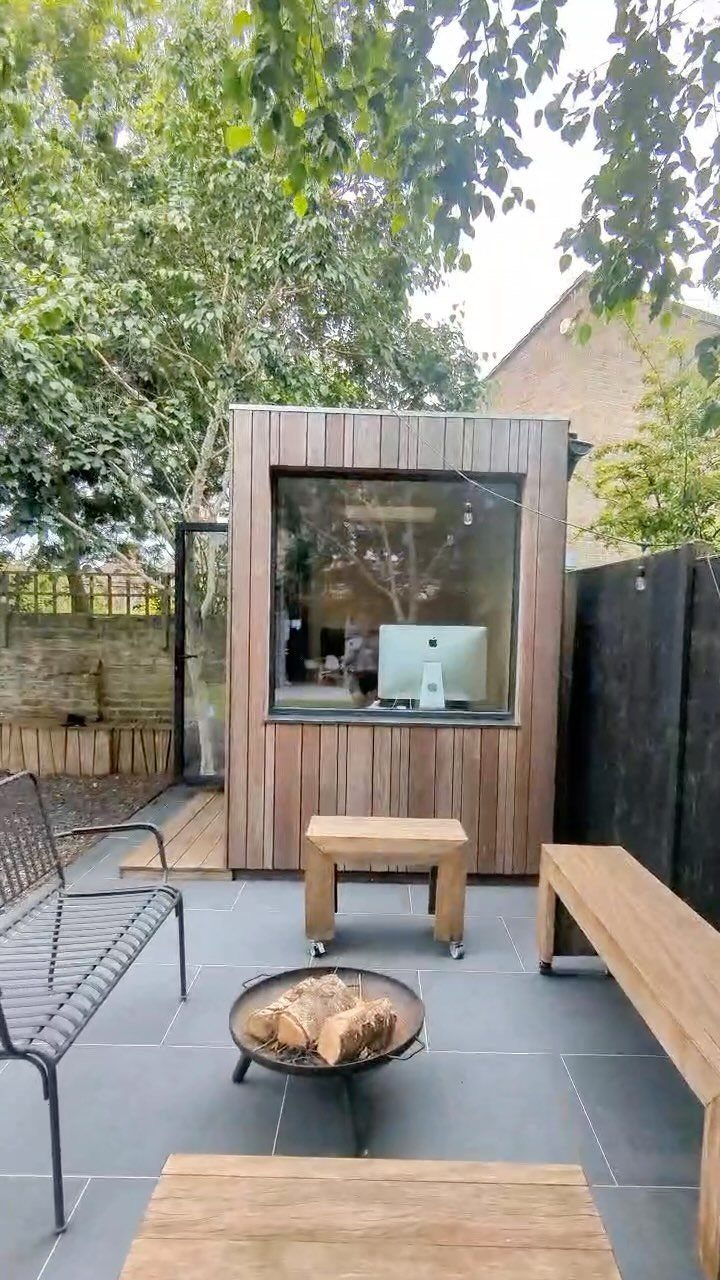 A modern wooden prefab studio with a large window overlooking a tiled backyard patio.