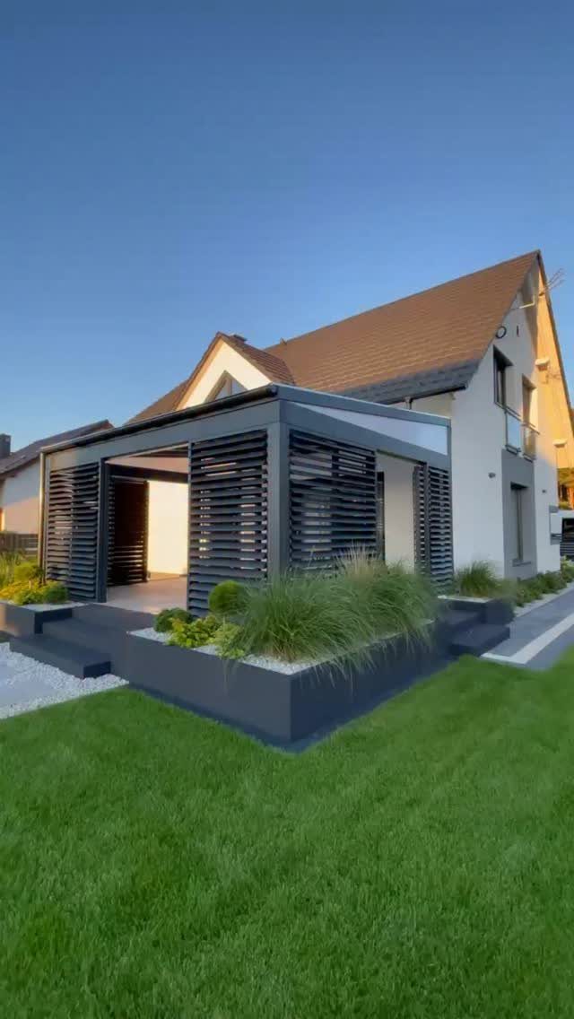 A modern dark grey pergola structure with slatted sides attached to a white house.