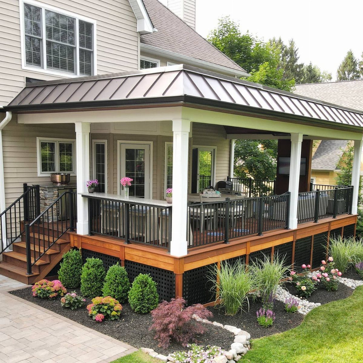 A wooden side house deck with a dark metal roof and white columns overlooking a garden.