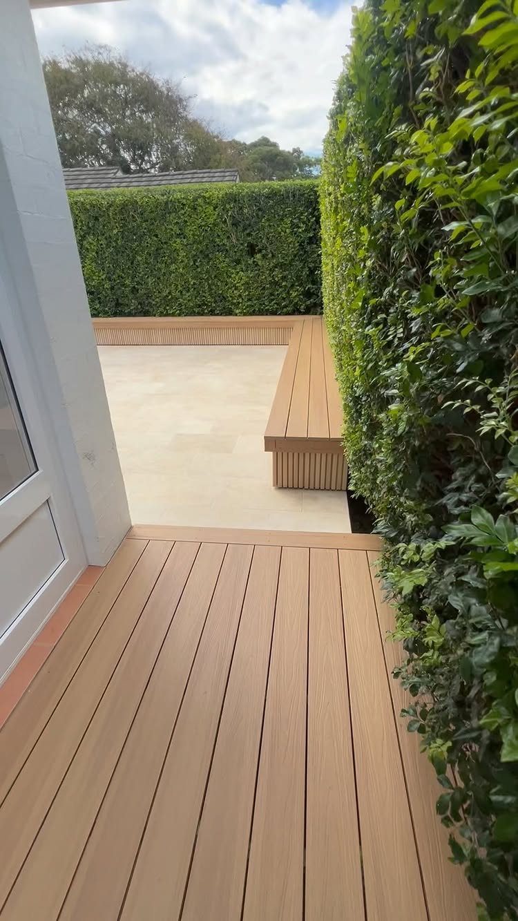 A wooden deck leads to a stone patio enclosed by tall green hedges.