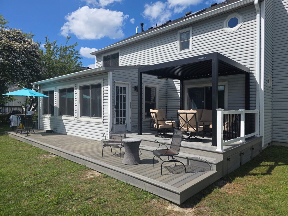 A grey composite deck with outdoor furniture sits adjacent to a light grey house and sunroom.