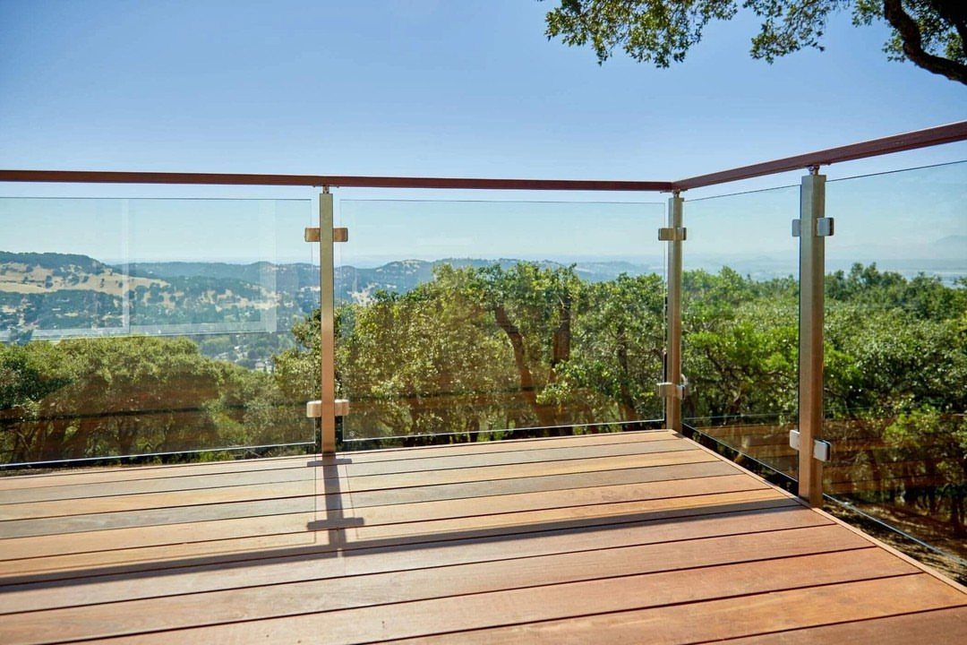 Wooden deck with transparent glass railings overlooking a scenic green valley and hills.
