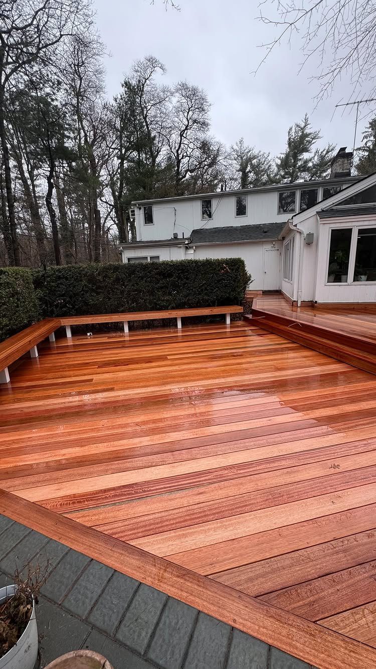 A polished reddish-brown wooden deck featuring built-in perimeter seating near a white house and green hedges.