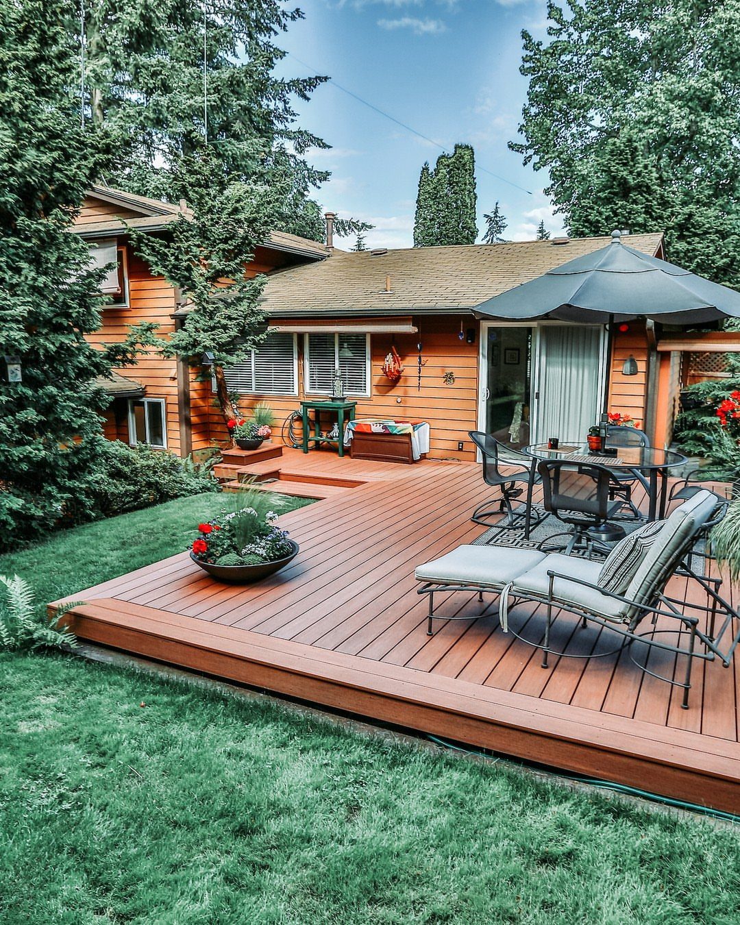 A low-profile wooden deck with outdoor furniture and a large black umbrella on a green lawn.