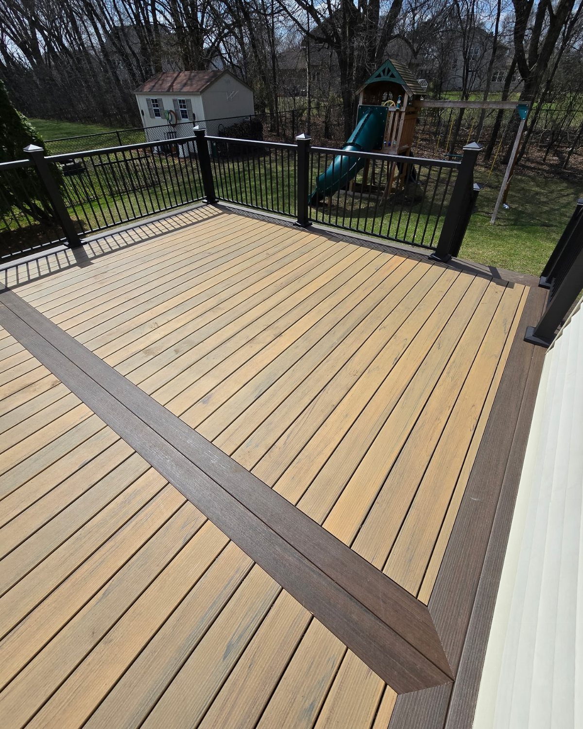 A wooden deck featuring a dark brown picture frame border and black metal railings in a backyard.