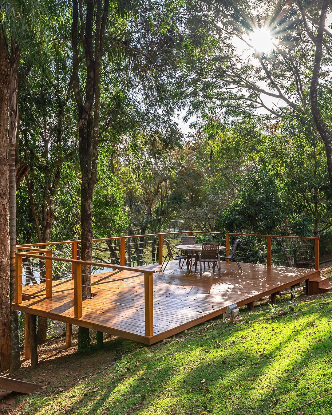 A wooden freestanding deck with cable railings situated on a grassy slope among tall green trees.