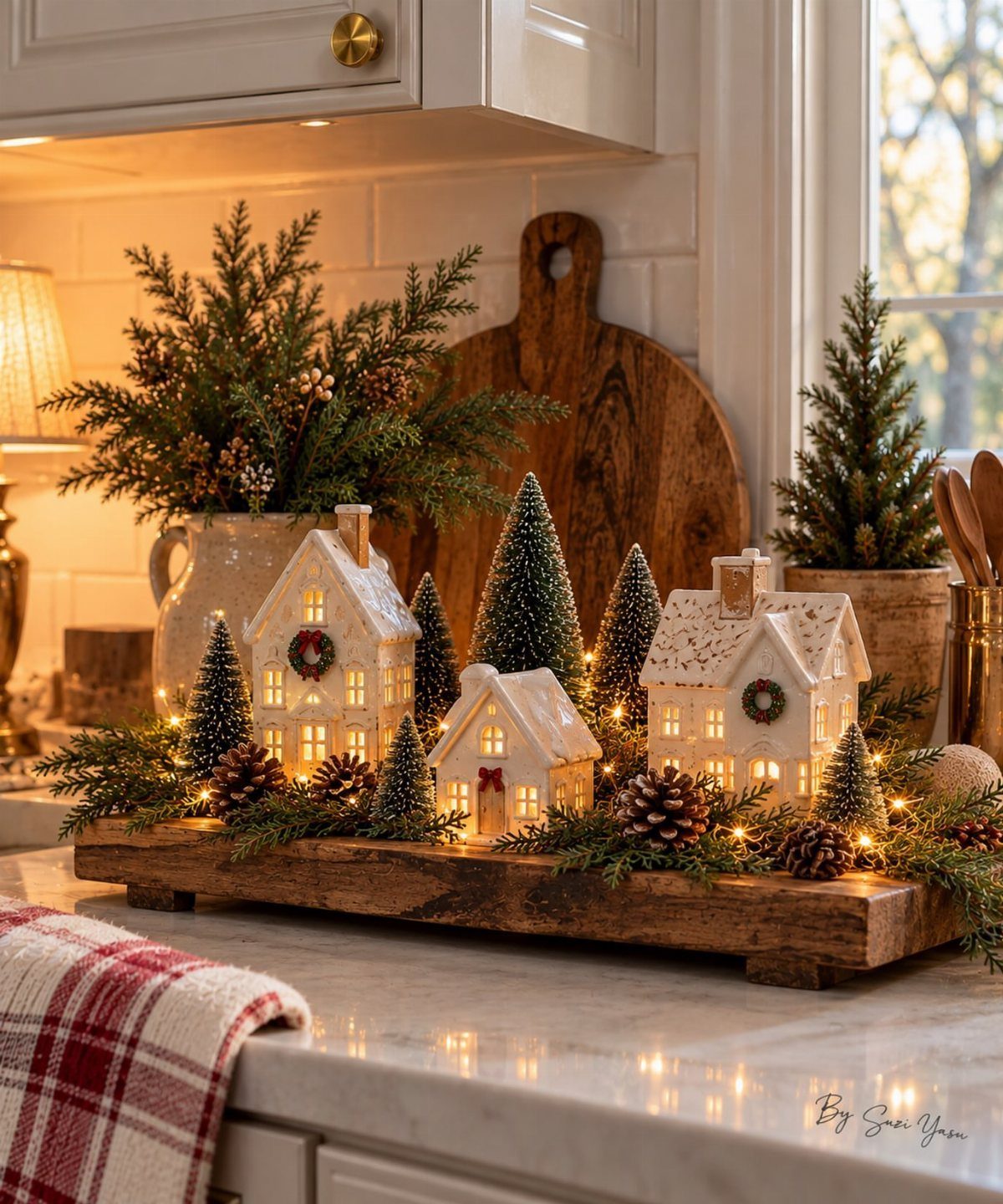 Miniature ceramic houses and small evergreen trees arranged on a wooden tray with lights.