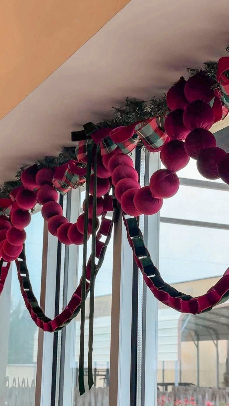 Red velvet pom-pom garland with plaid ribbon hanging across a window frame.